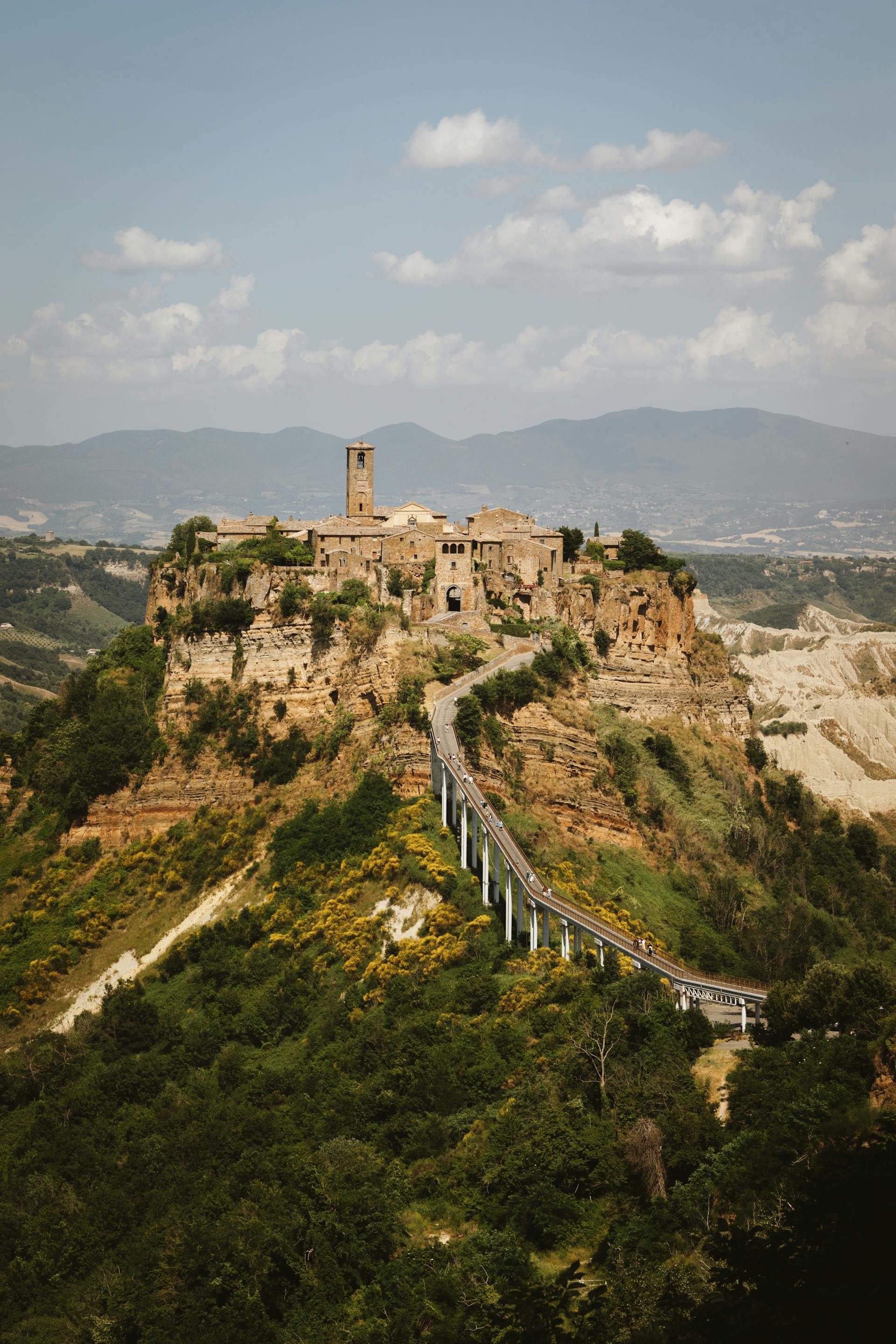 A historic village with stone buildings and a prominent bell tower, perched dramatically on a steep, eroding hilltop, accessed by a long, elevated pedestrian bridge.