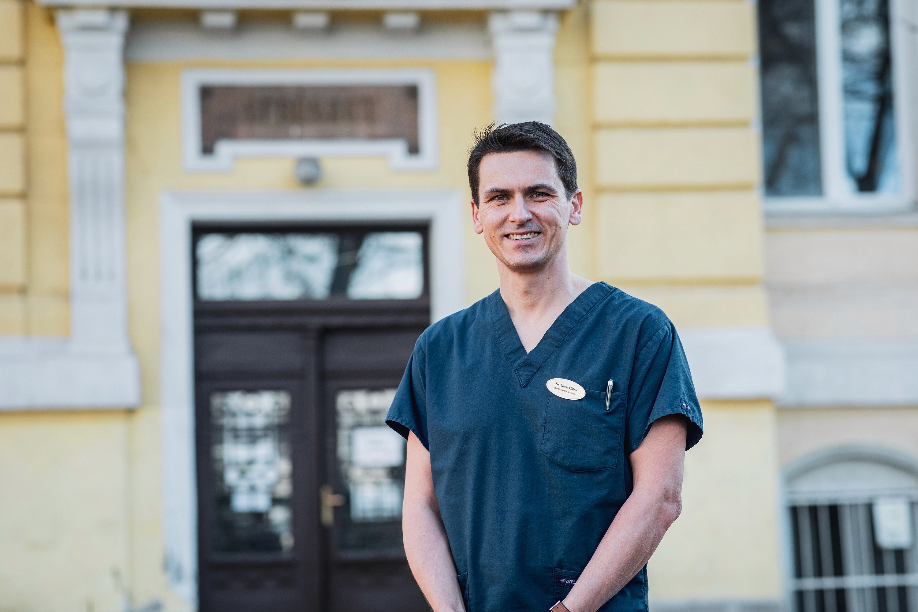 Mosolygós férfi orvos, sötétkék egyenruhában sárga kórházi épület előtt.