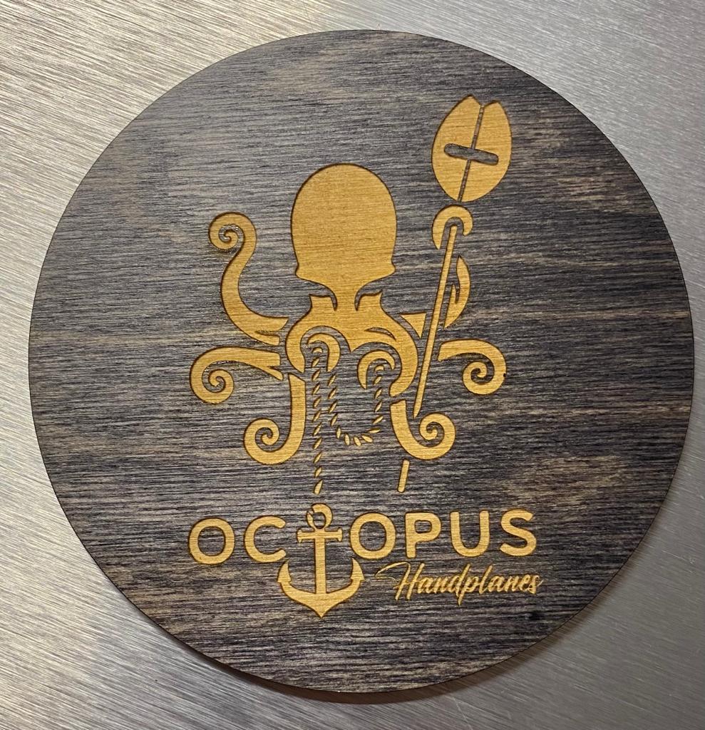 Sous-verre en bois avec une pieuvre gravée tenant une pagaie et une ancre. Le texte « Octopus Handplanes » est inscrit dans une police stylisée sur une surface texturée.