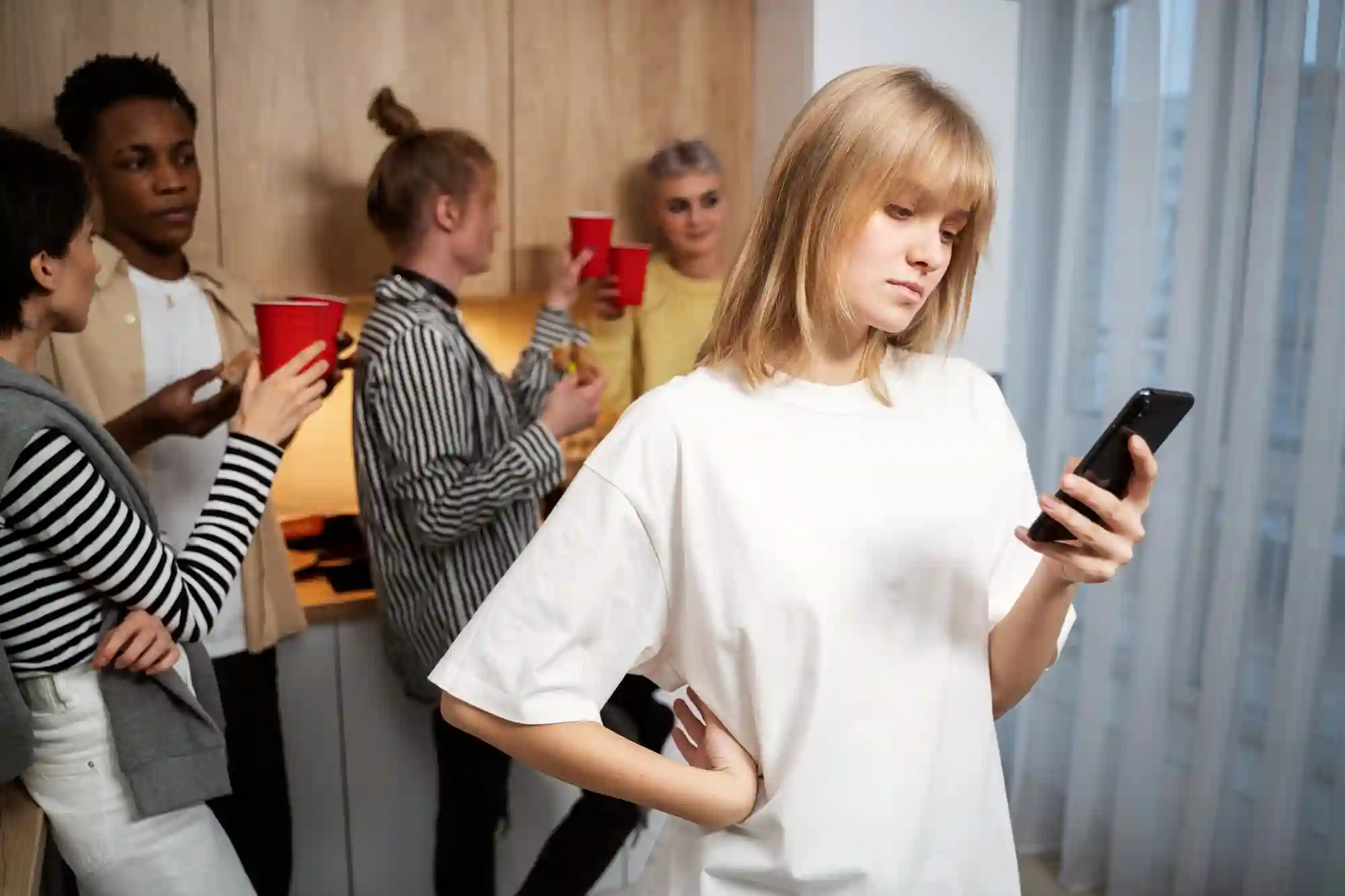 A woman looking at her phone while feeling socially isolated at a busy house party with friends nearby.