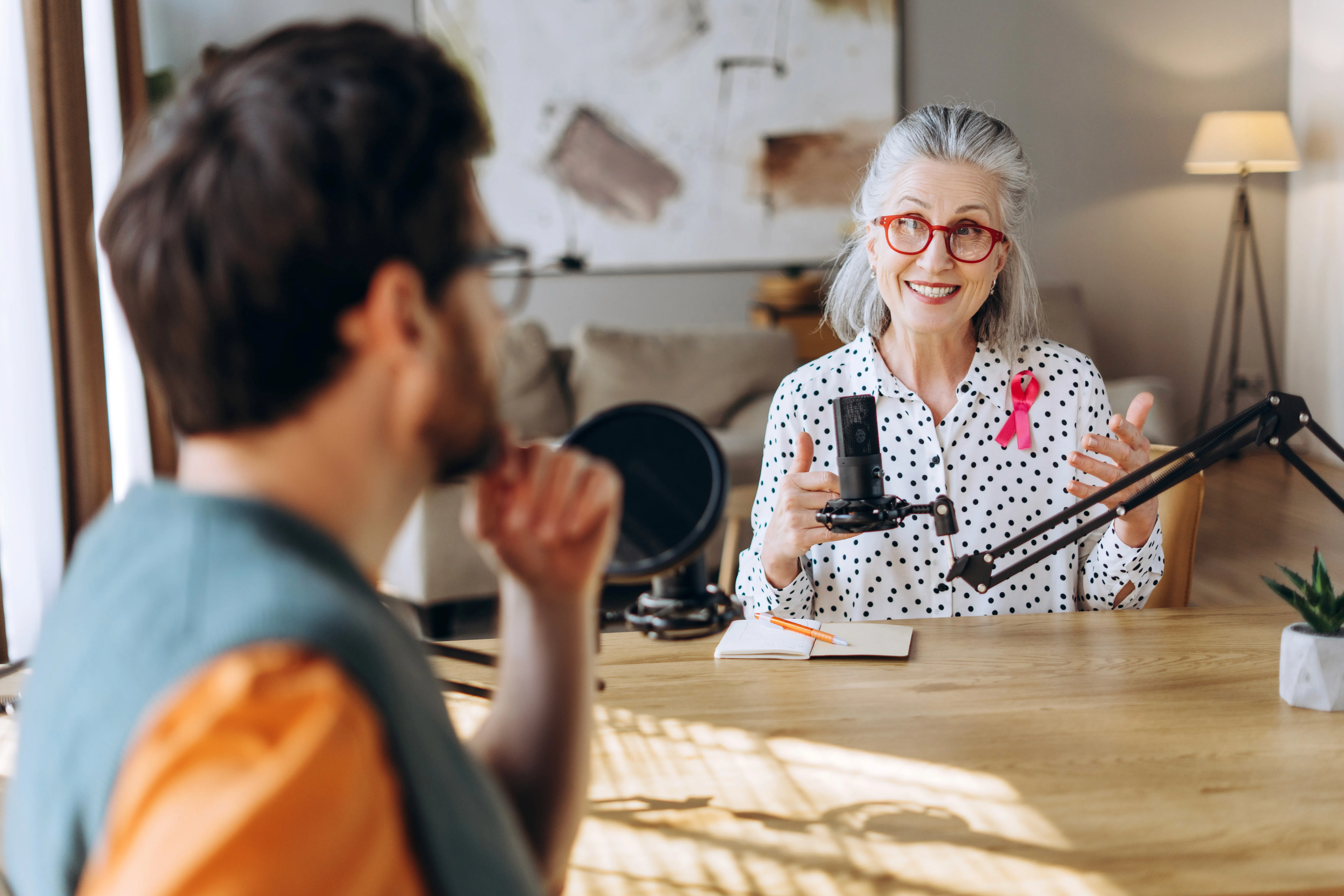 Man wearing an olive green jacket and black shirt, sitting and smiling in front of a light wooden background.