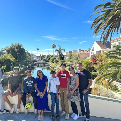 Grupo de nueve personas posando en un puente con un canal pintoresco, palmeras y casas de fondo bajo un cielo azul claro.