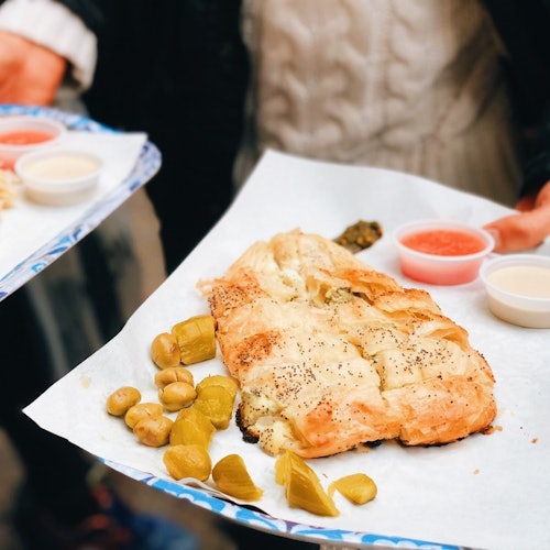 A tray with a pastry, pickles, and sauces. Two hands holding the tray, wearing sweaters.