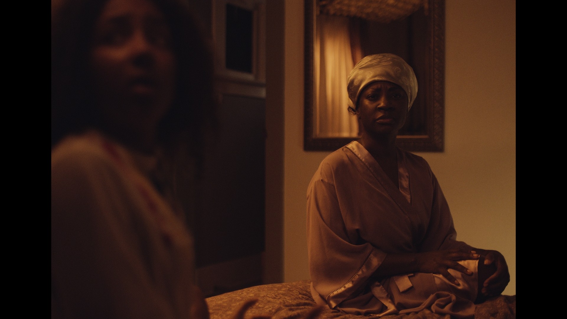 One dark-skinned black woman (Natasha Ofili as Victoria) and one black girl (Mila Davis-Kent as Sarafina) in a dimly lit room. Victoria is seated on a bed wearing a satin headscarf and robe, and Sarafina is blurred in the foreground.