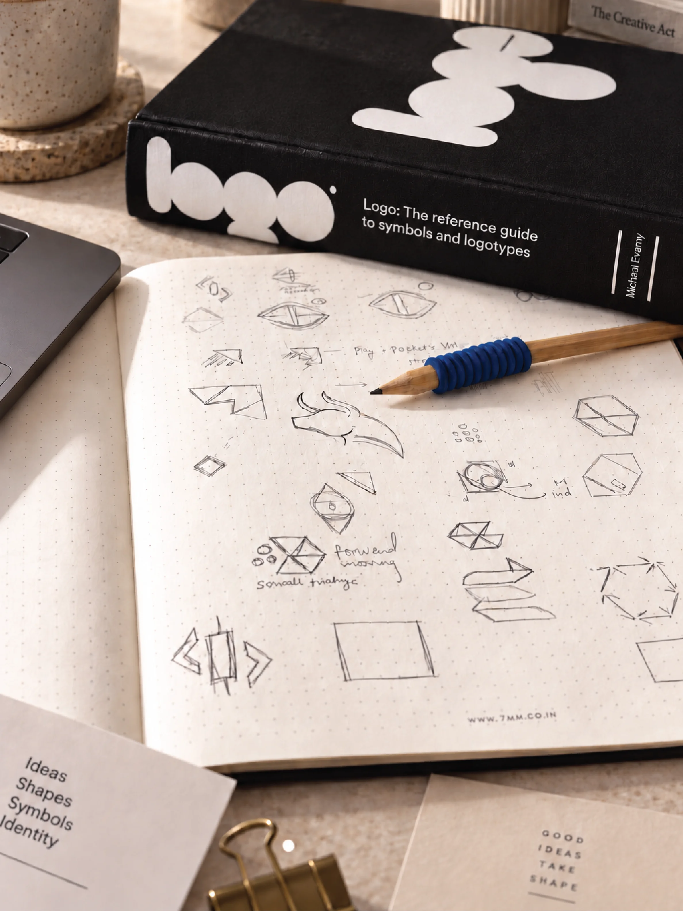 An open sketchbook filled with logo design sketches in pencil, featuring geometric shapes and symbols, is placed on a table next to a branded book titled "Logo: The reference guide to symbols and logotypes," accompanied by a pencil with a blue grip, a laptop, and a paper clip; perfect for creative design and branding inspiration.