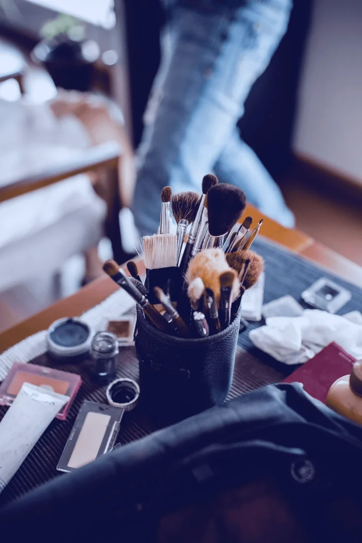 A collection of assorted makeup brushes and tools is neatly arranged in a black holder on a wooden vanity table, surrounded by various cosmetics.