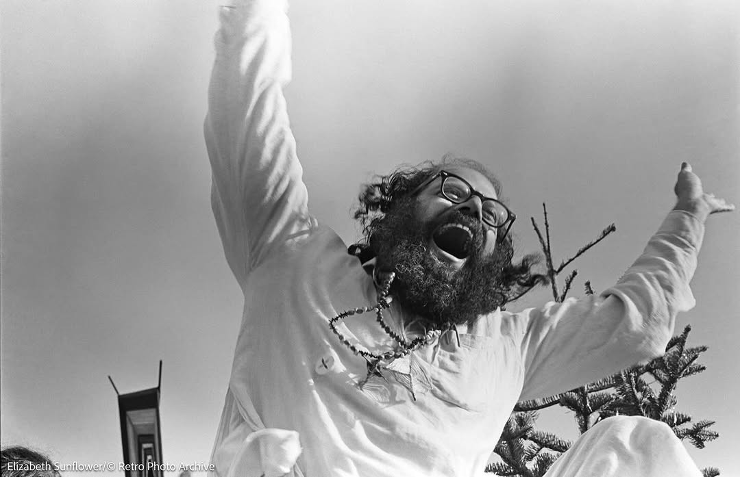 Black-and-white photograph of a bearded man in white laughing with both arms raised.