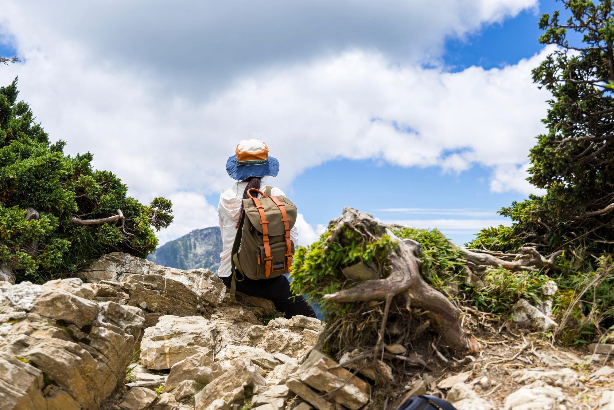 A back view of a female hiker sitting on a rocky mountain ledge, wearing a wide-brimmed sun hat and a green canvas backpack with leather straps, looking out at a mountain peak under a cloudy blue sky.