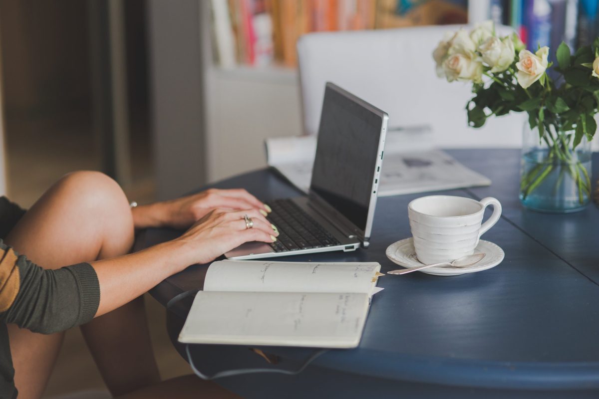 A person types on a laptop while sitting awkwardly at their desk with one knee up.