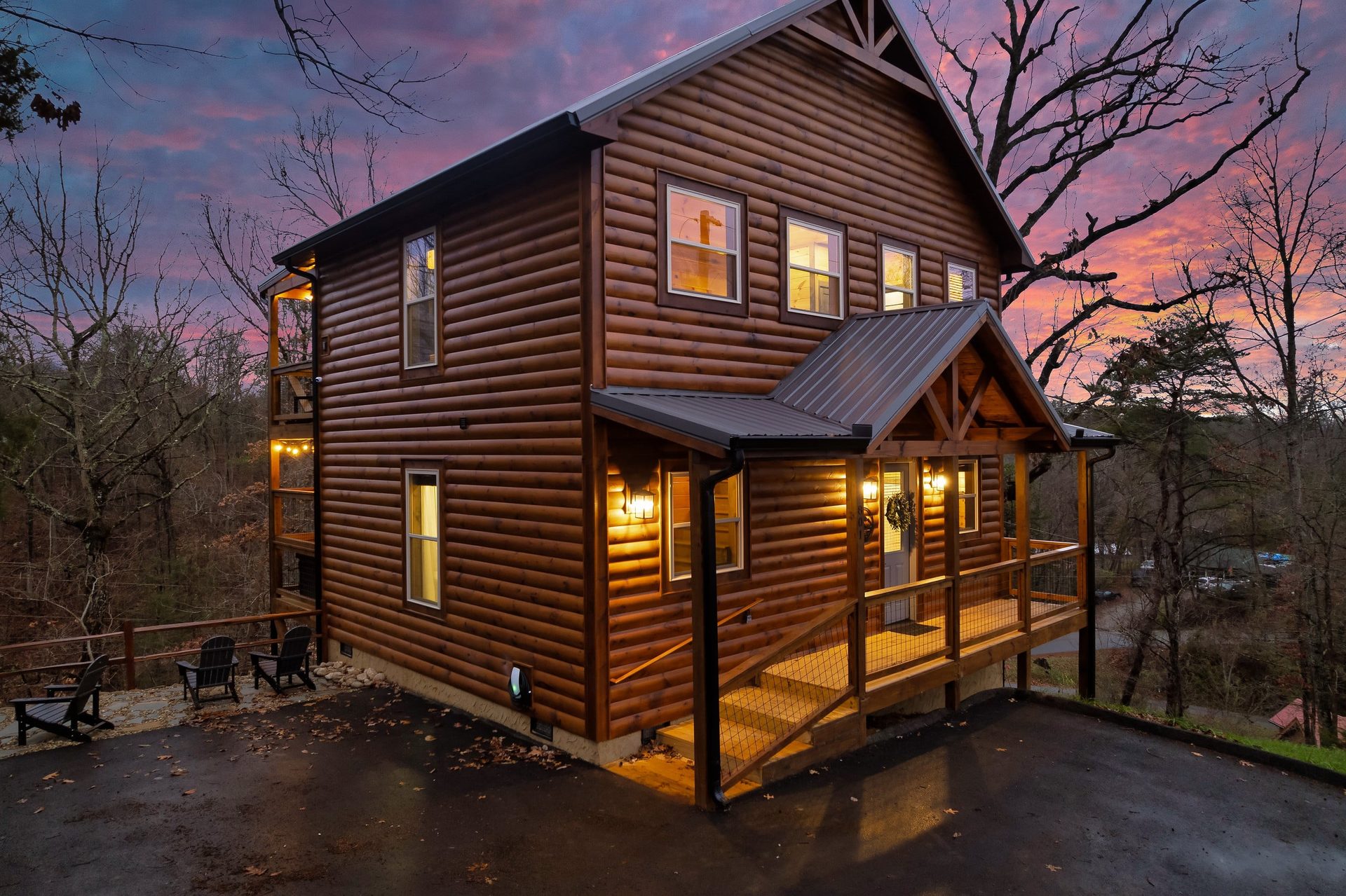 Front of the log cabin at dusk with a covered entry porch, paved parking area and Adirondack chairs at the fire pit