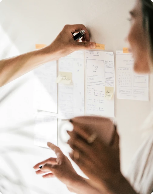 A close-up of a collaborative brainstorming session where a person is taping paper wireframe sketches for a mobile app onto a white wall