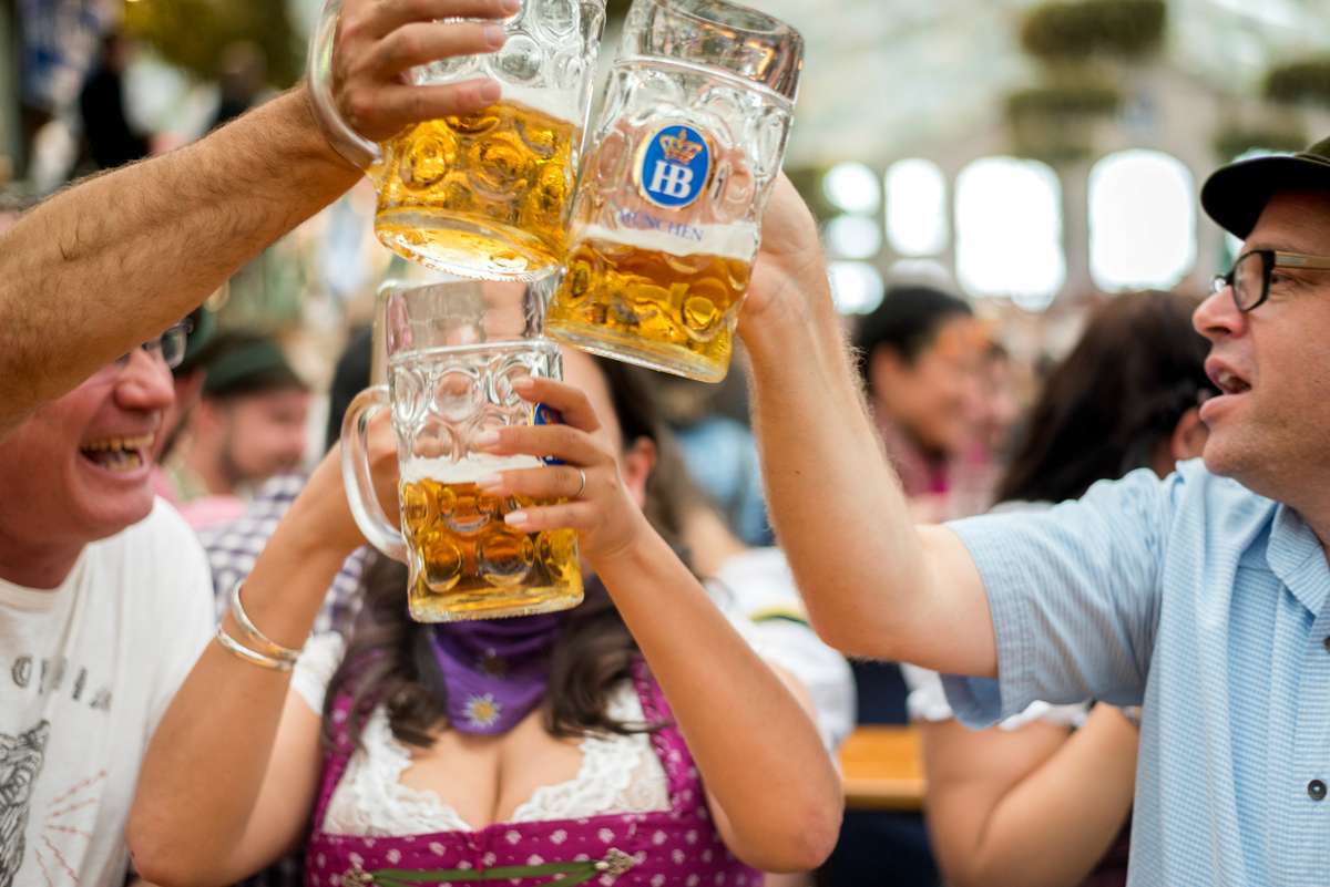 People in traditional Bavarian attire clink large glass beer mugs filled with golden beer and white foam at a festival.
