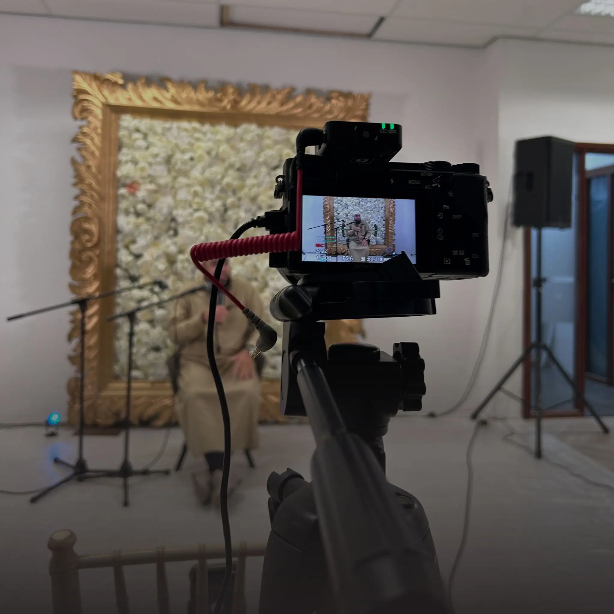 Een cameraman filmt een persoon die op een stoel zit in een studio, met een gouden lijst om een bloemenachtergrond en microfoons in de buurt.