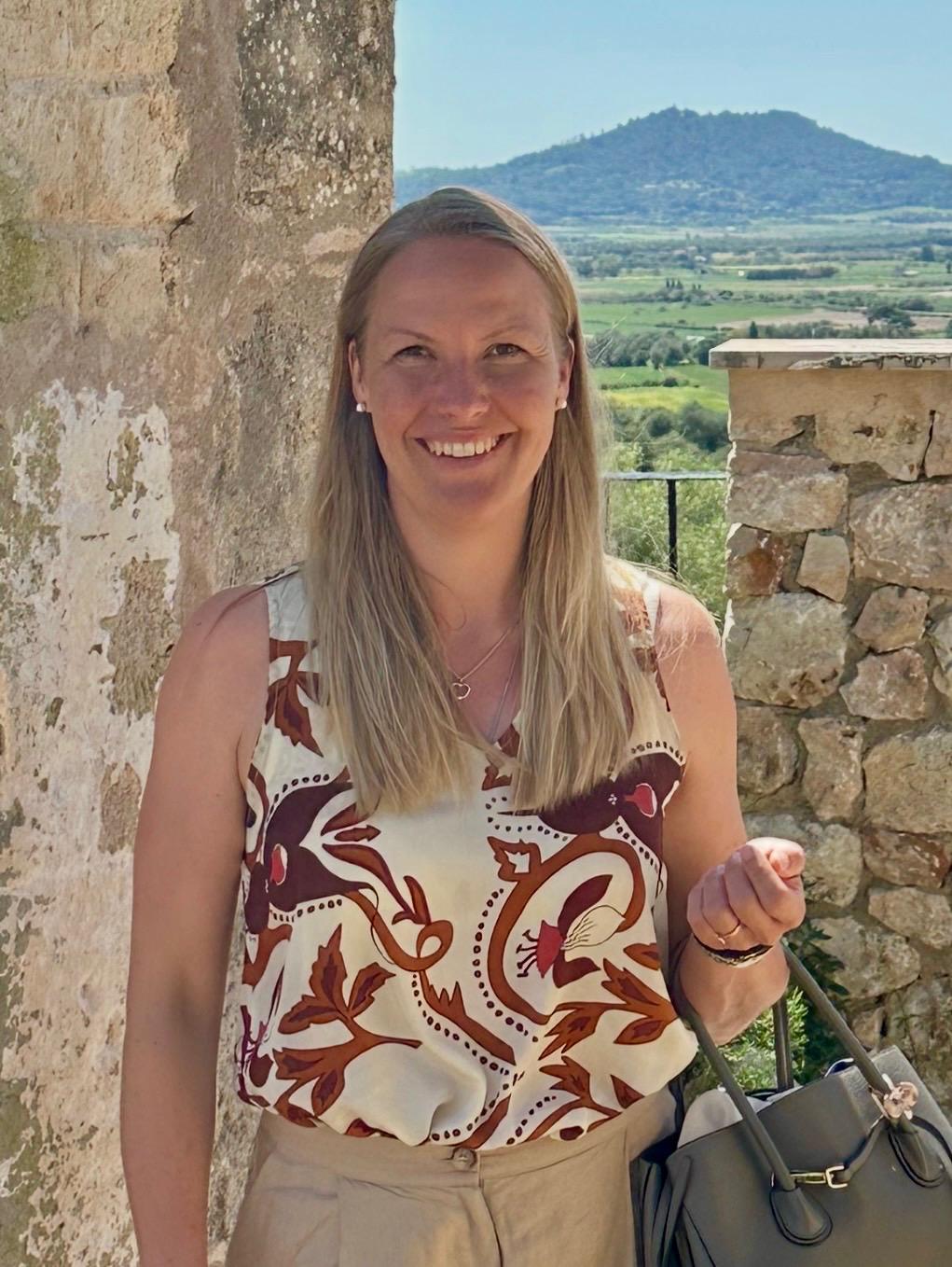 Black-and-white portrait of a young man with short hair and light facial stubble, looking directly at the camera with a neutral expression, outdoors with blurred buildings and trees in the background.