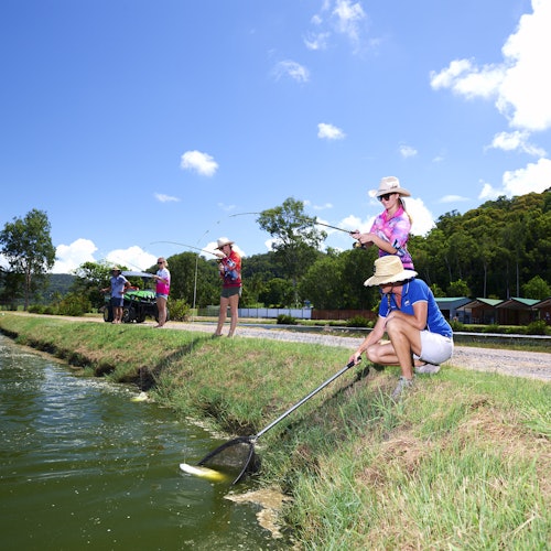 一群人在晴朗的蓝天下池塘边钓鱼。一个人跪着拿着网，其他人则站着拿着钓鱼竿。