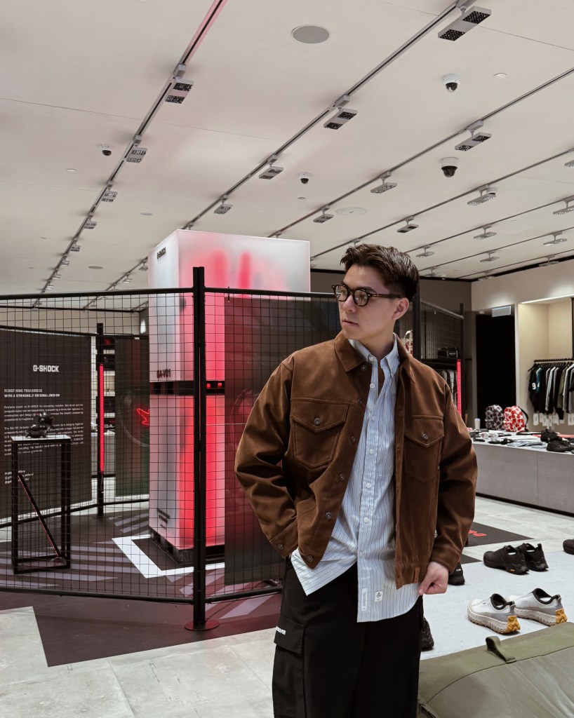 A young man wearing a brown jacket over a blue shirt stands in a modern retail store with low lighting and a stylish, contemporary design.