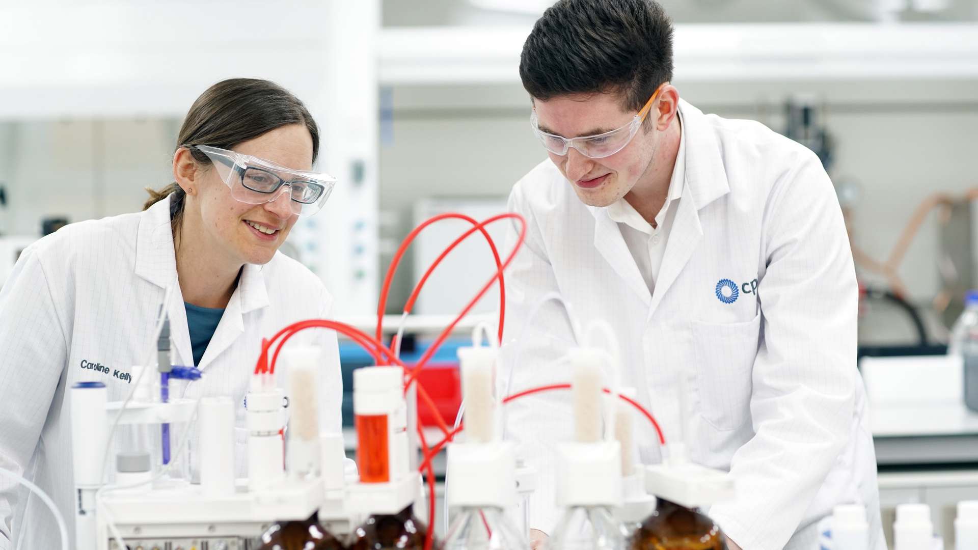 Two CPI scientists in white lab coats and safety glasses smiling as they examine amber bottles and titration equipment with red tubing