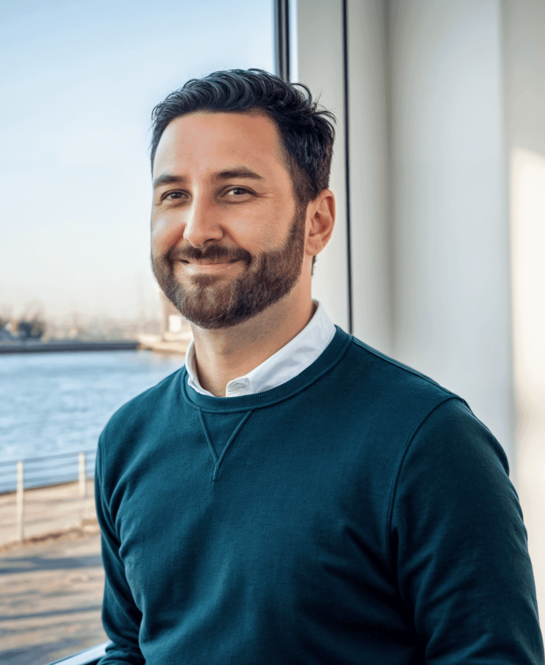 A man with a beard wearing a green sweater stands in front of a window, gazing outside.