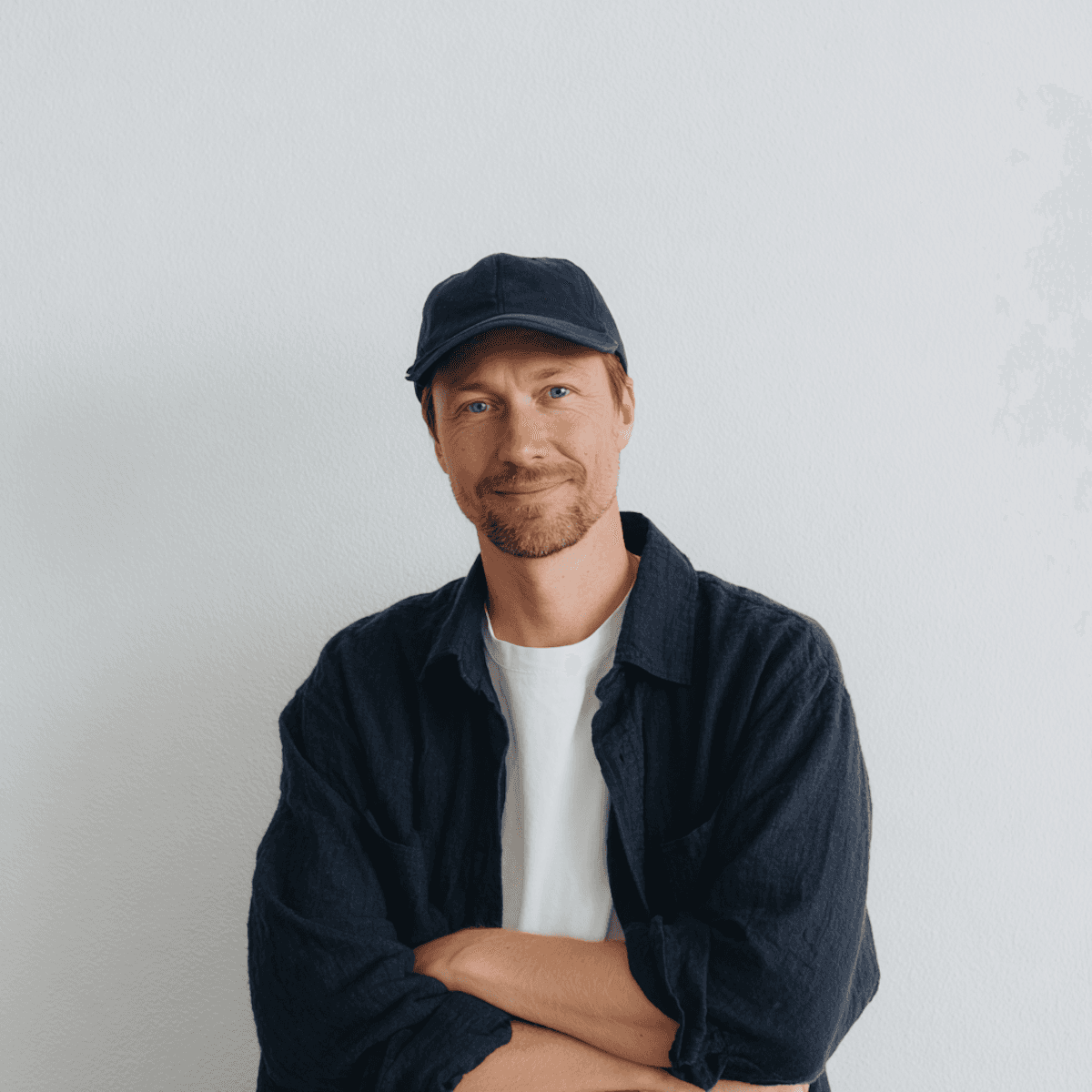 Man wearing a dark cap, white T-shirt, and navy overshirt, standing with arms crossed and smiling softly in front of a plain light-colored wall.
