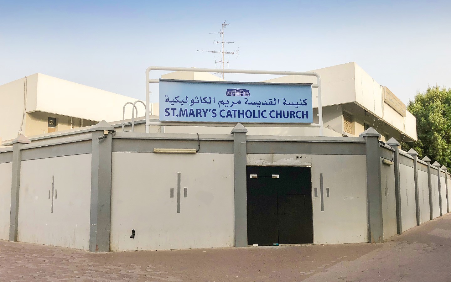 The white-walled exterior and black gate of St. Mary's Catholic Church, featuring a sign with the church name in English and Arabic.