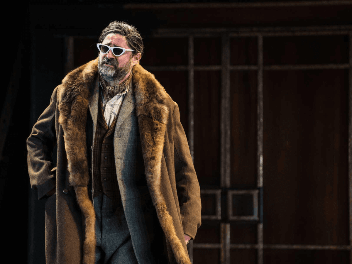 Man in a fur coat and sunglasses posing in a stylish Twelfth Night scene.