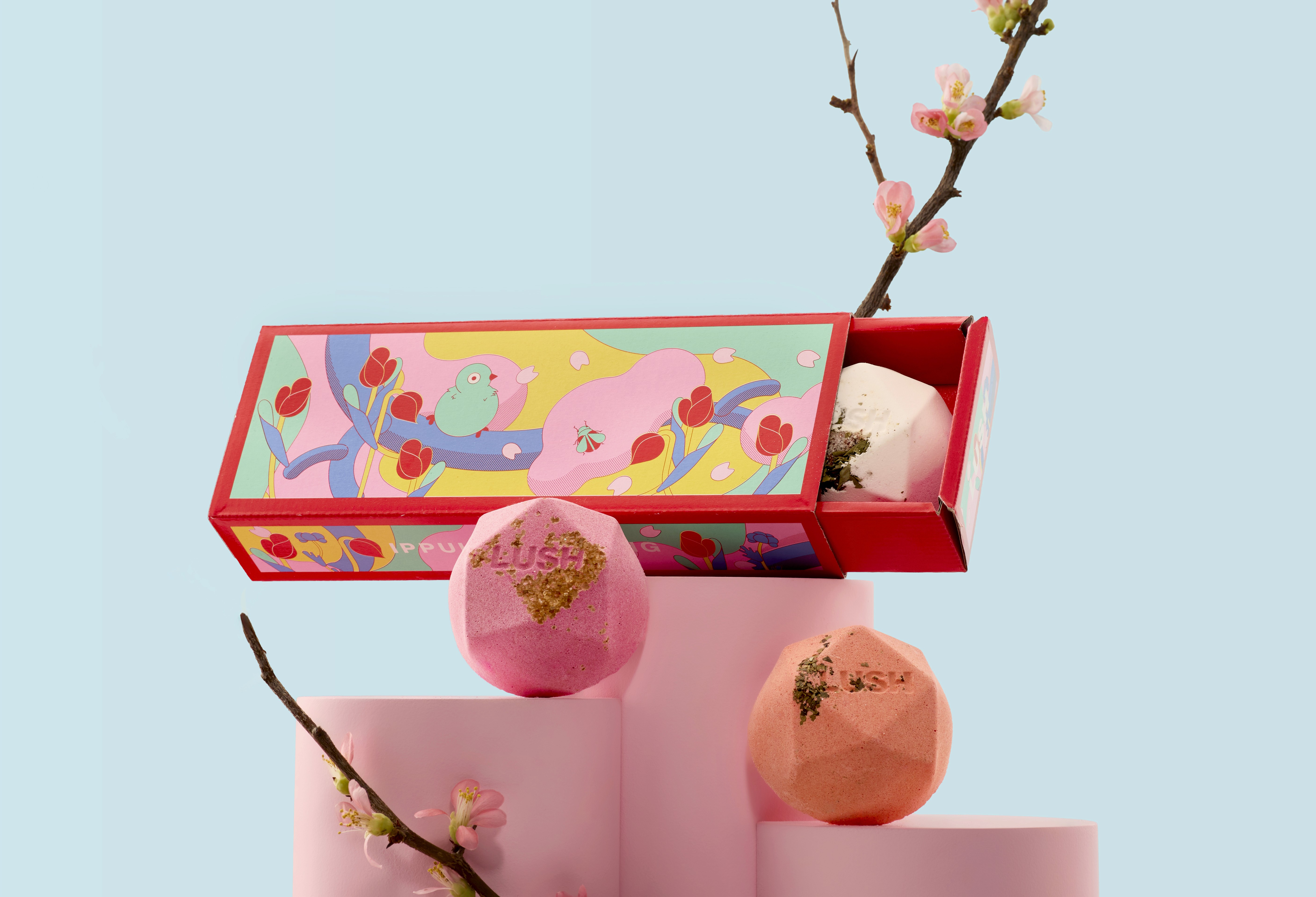 Rectangular Lush gift box with colorful spring illustration, partially opened to reveal bath bombs, displayed on pink blocks with cherry blossom branches.