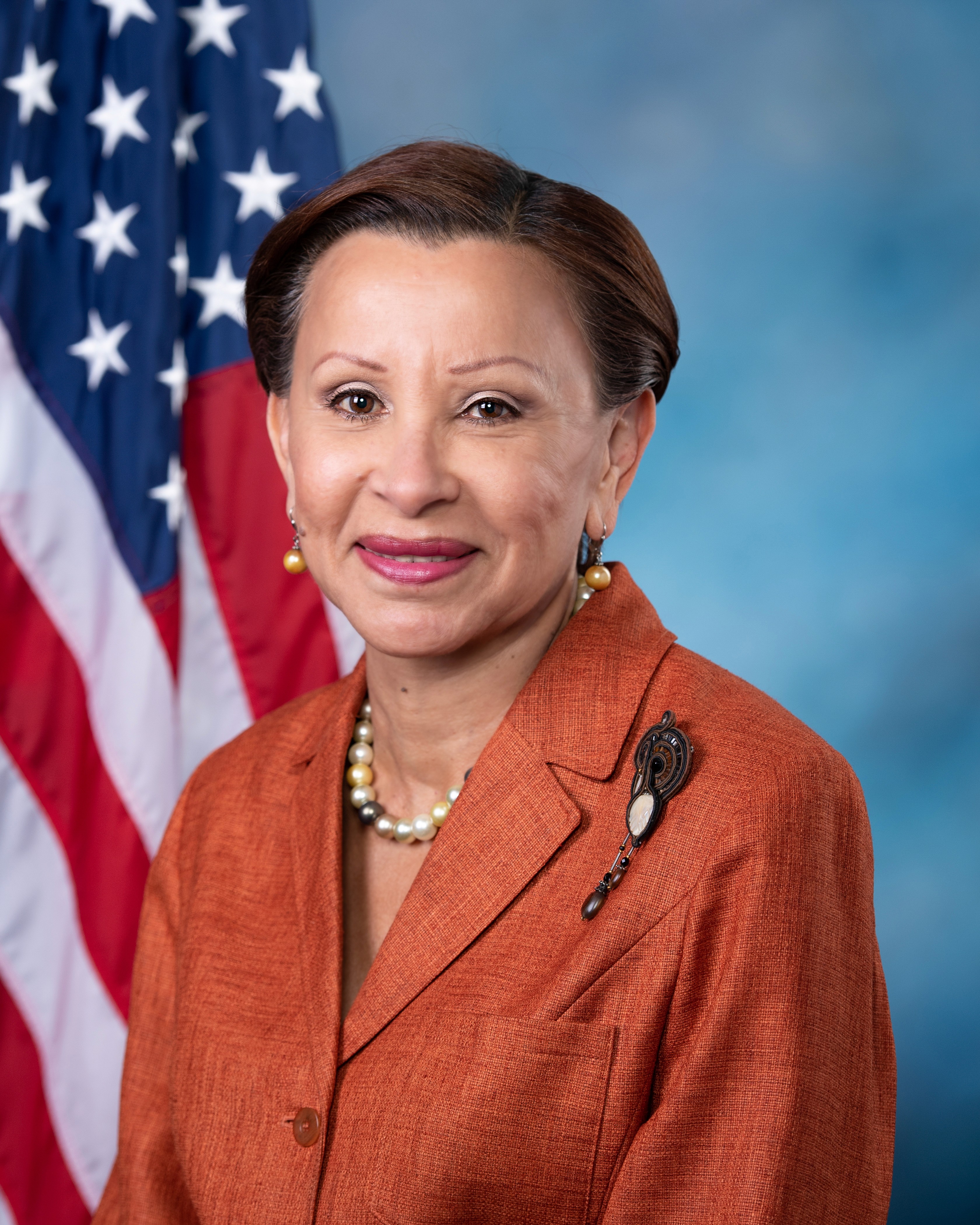 Portrait of Nydia M. Velázquez on a blue background with a portion of the flag of the United States of America in view behind subject