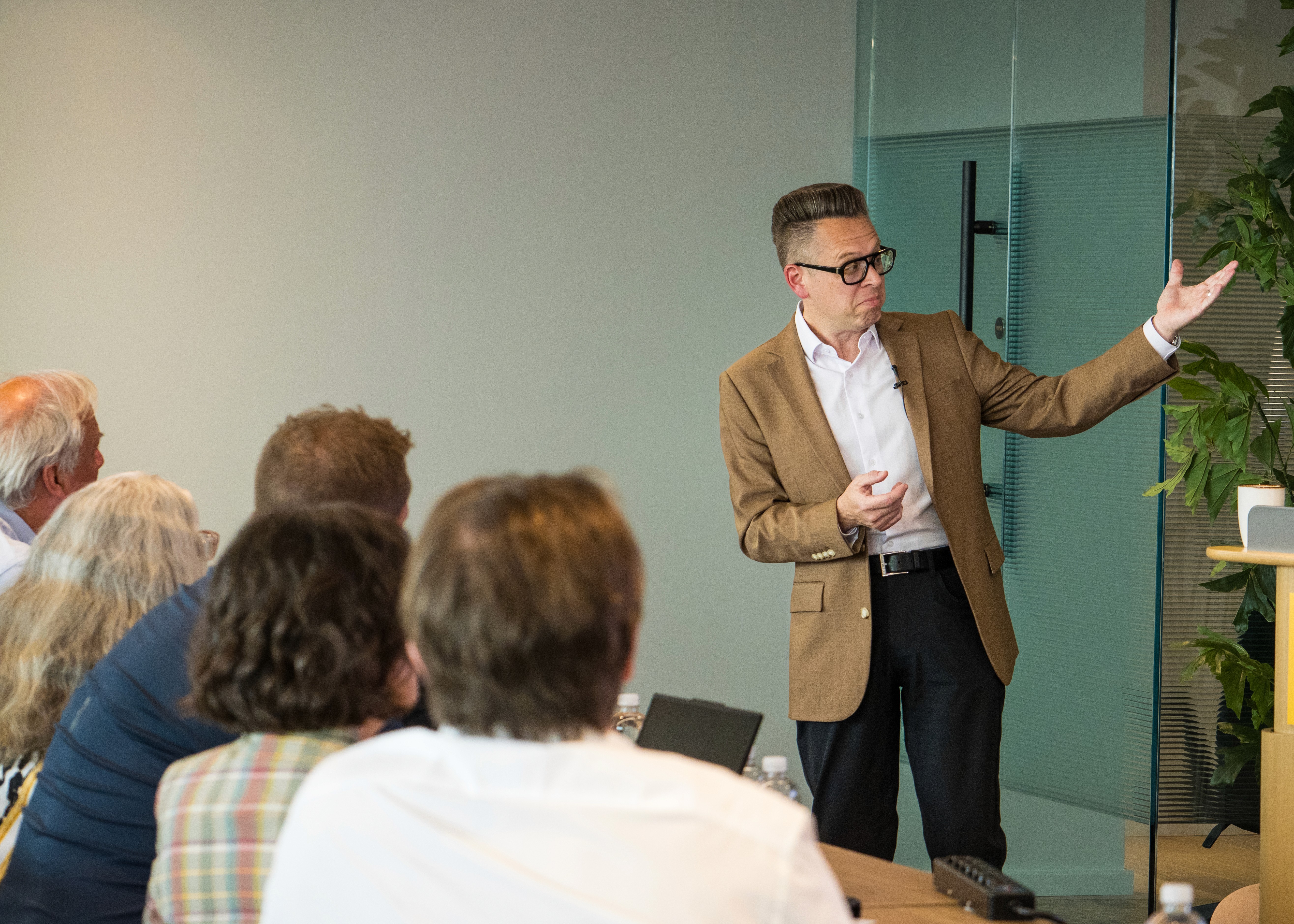 A medium shot of a presentation in progress inside a professional corporate or conference setting. On the right, a man with short, styled dark hair and black-rimmed glasses is speaking while wearing a brown tweed blazer, a white button-down shirt, a black belt, and dark trousers. He is gesturing with his left arm extended outward toward a glass wall and a tall potted plant in the background. On the left side of the frame, several audience members are seated at a table, viewed from behind or in profile, listening to the speaker.