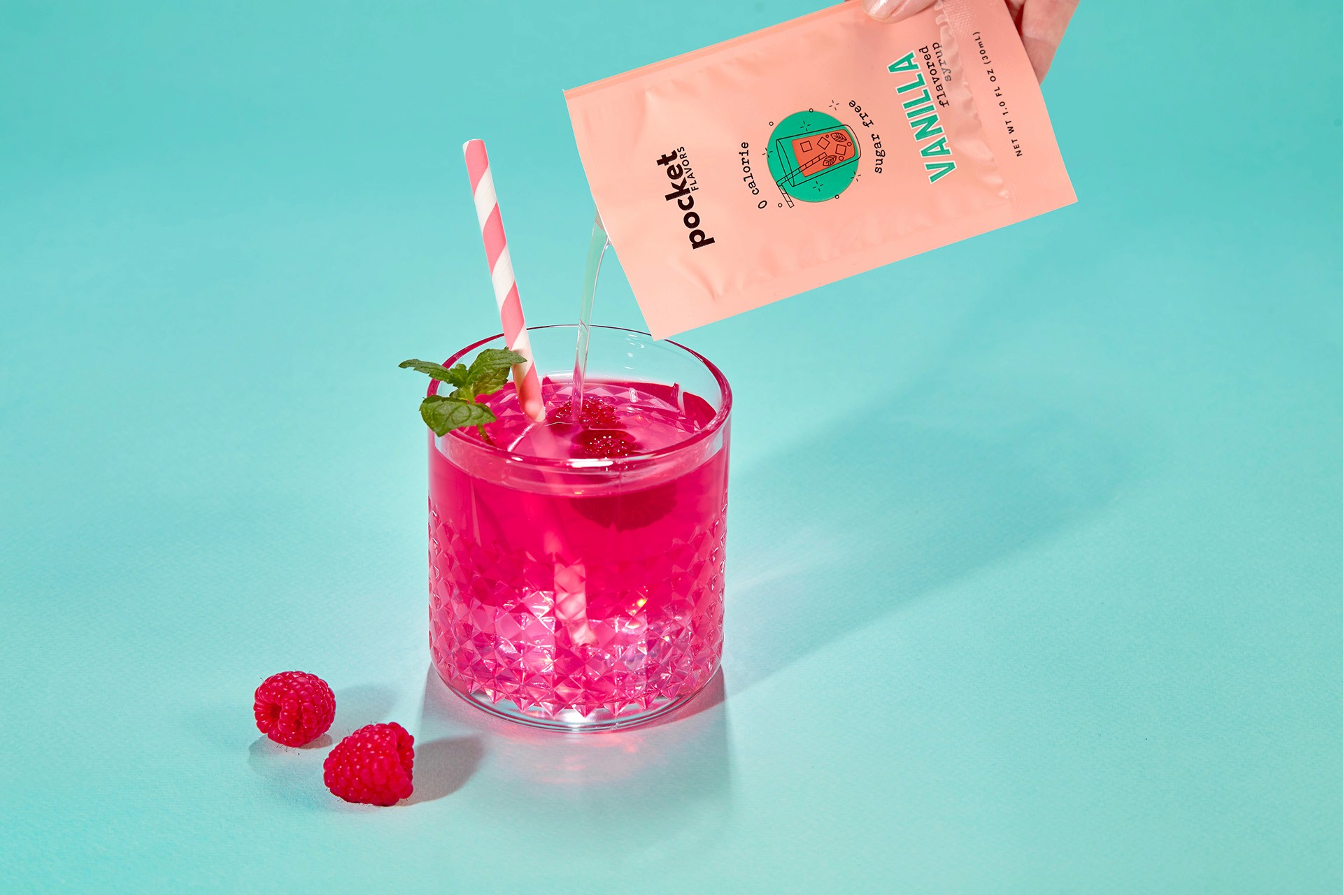 A hand pours a Packet of Pocket Flavors vanilla mix into a glass of vibrant pink raspberry drink, garnished with fresh mint and accompanied by a striped straw, set against a turquoise background.