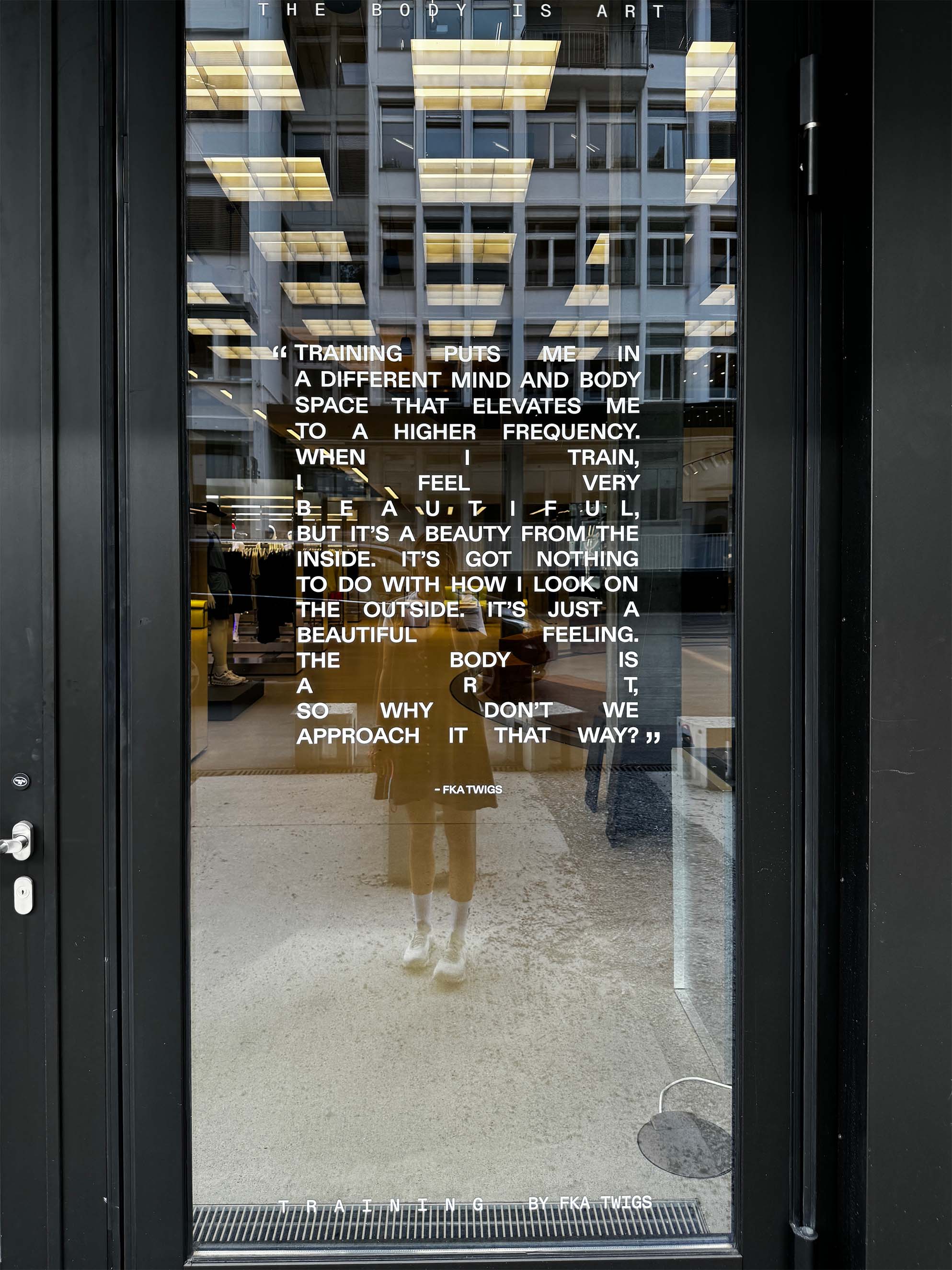 A glass door with white text displaying a motivational quote about training, beauty, and the body. Reflections of a city street with cars and buildings hint at the creative studio inside, inspiring every campaign and strategy beyond the glass.