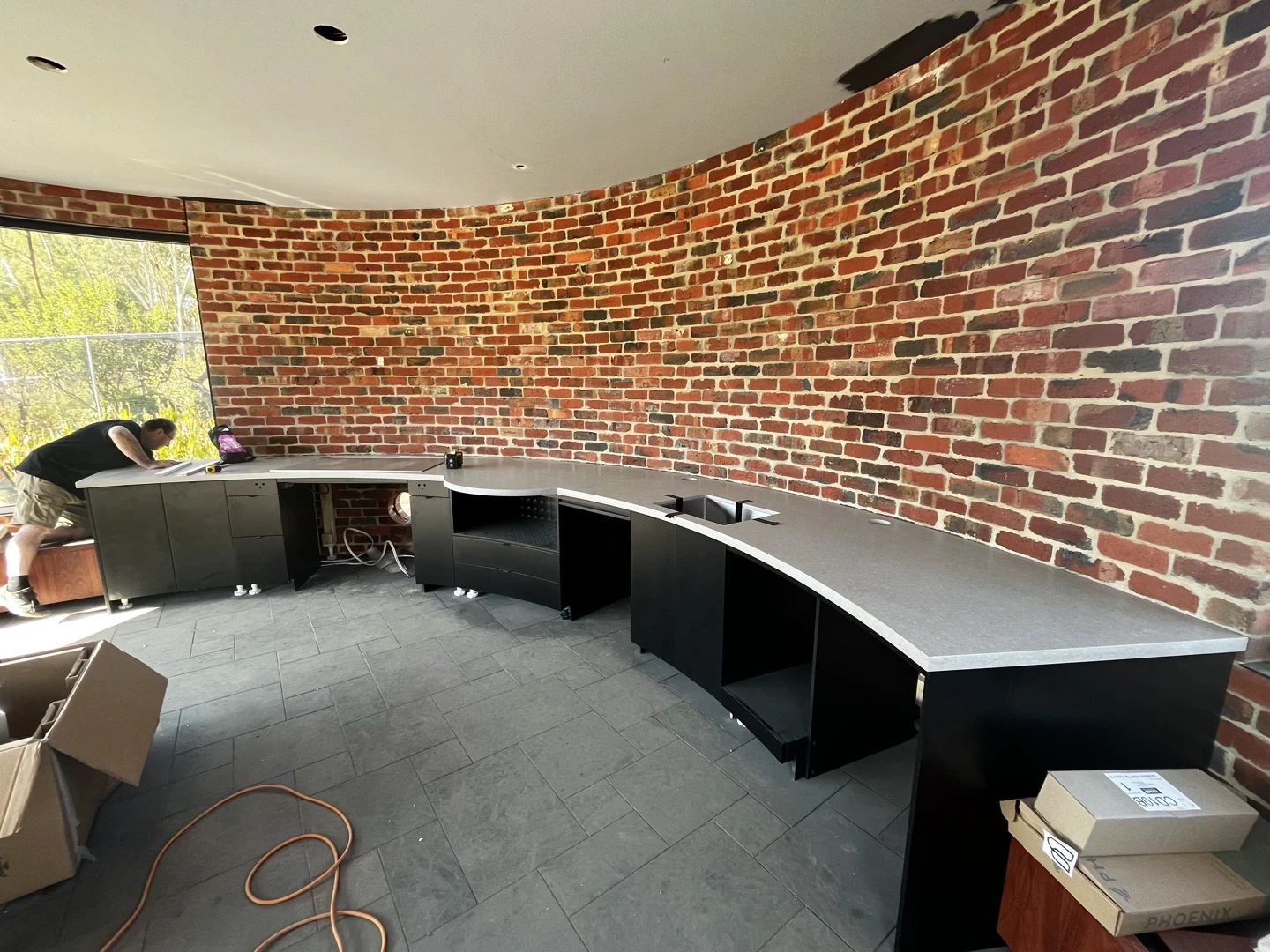 Curved natural stone commercial counter with dark work surface, brick wall backdrop, and fitted joinery.