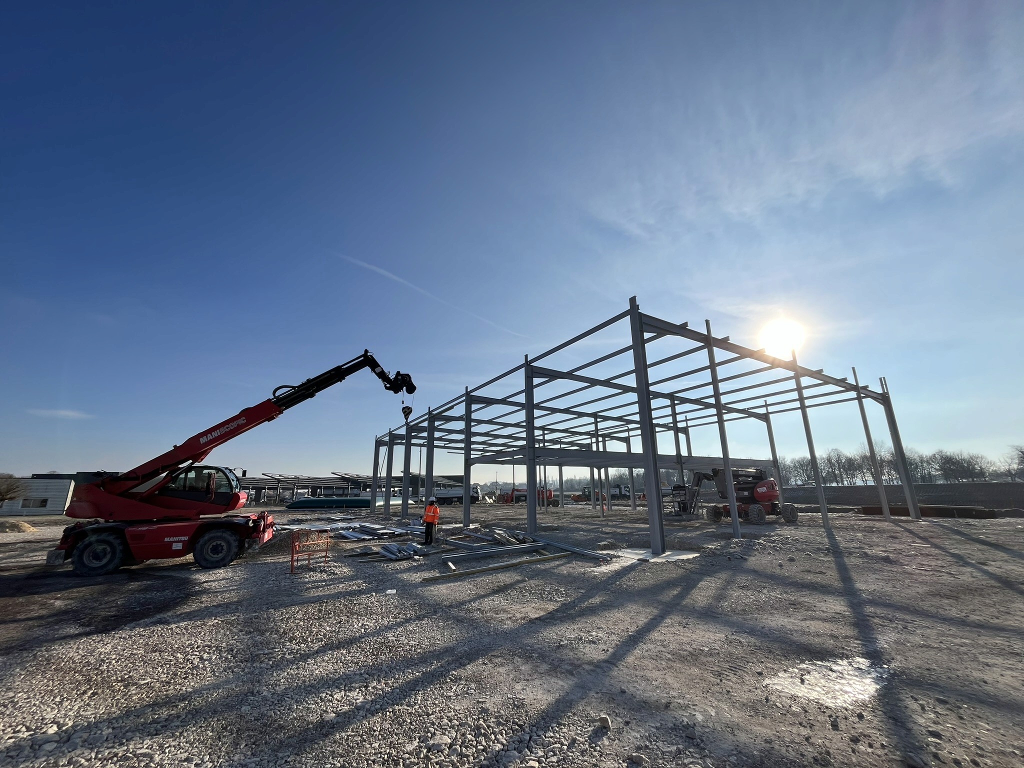 Montage d’une structure métallique avec grue rouge sous un ciel bleu et ensoleillé, sur un terrain de chantier dégagé.