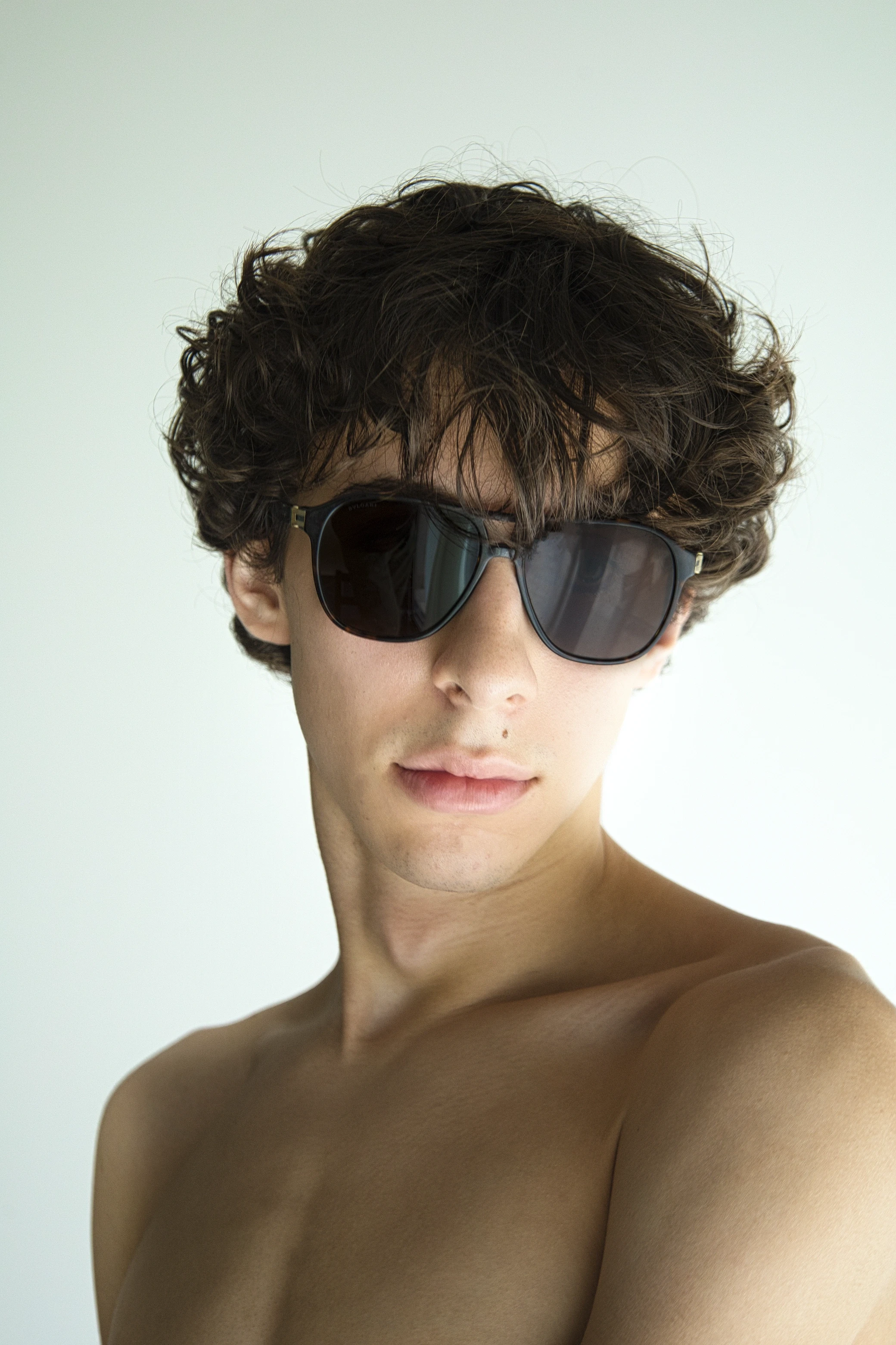 A young person with curly hair wearing black sunglasses, facing slightly away against a light background.