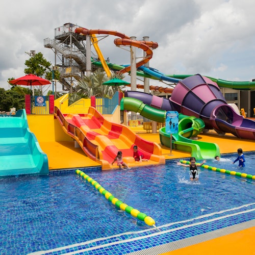 Children playing at a colorful water park with multiple slides and a pool, under a cloudy sky.