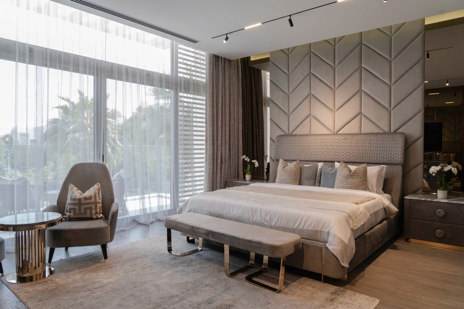 A modern, luxurious bedroom features a spacious king-sized bed with a textured gray headboard, complemented by matching bedding and pillows, set against a backdrop of large windows draped with sheer curtains, allowing natural light to illuminate the room, which includes an elegant armchair and a sleek side table with decorative items.