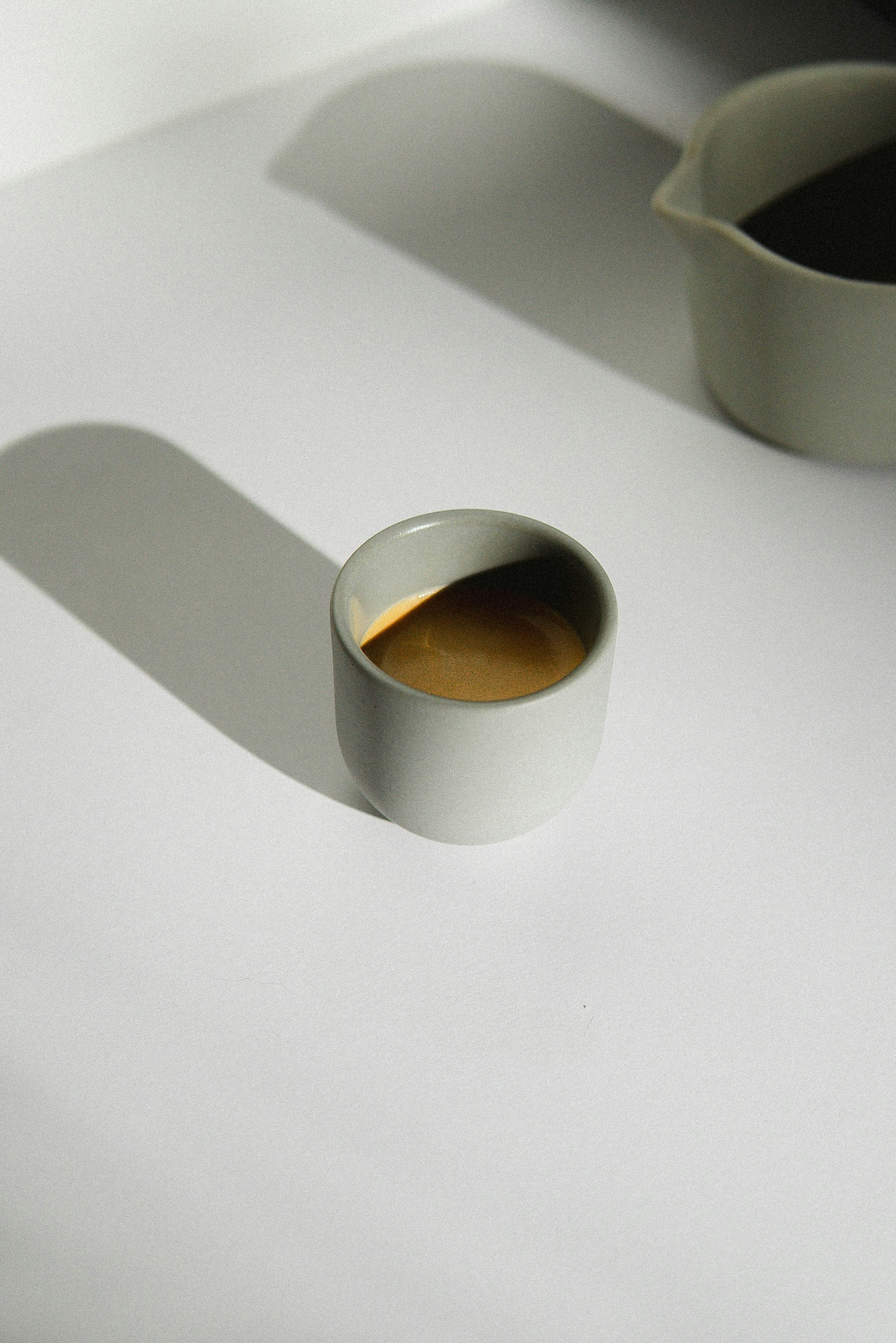 A round, white ceramic cup with brown coffee inside, casting a shadow on a white surface.