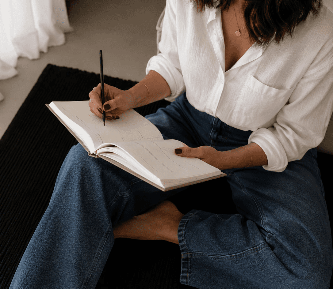 Woman sitting indoors writing in a notebook with a pen, looking to the side.
