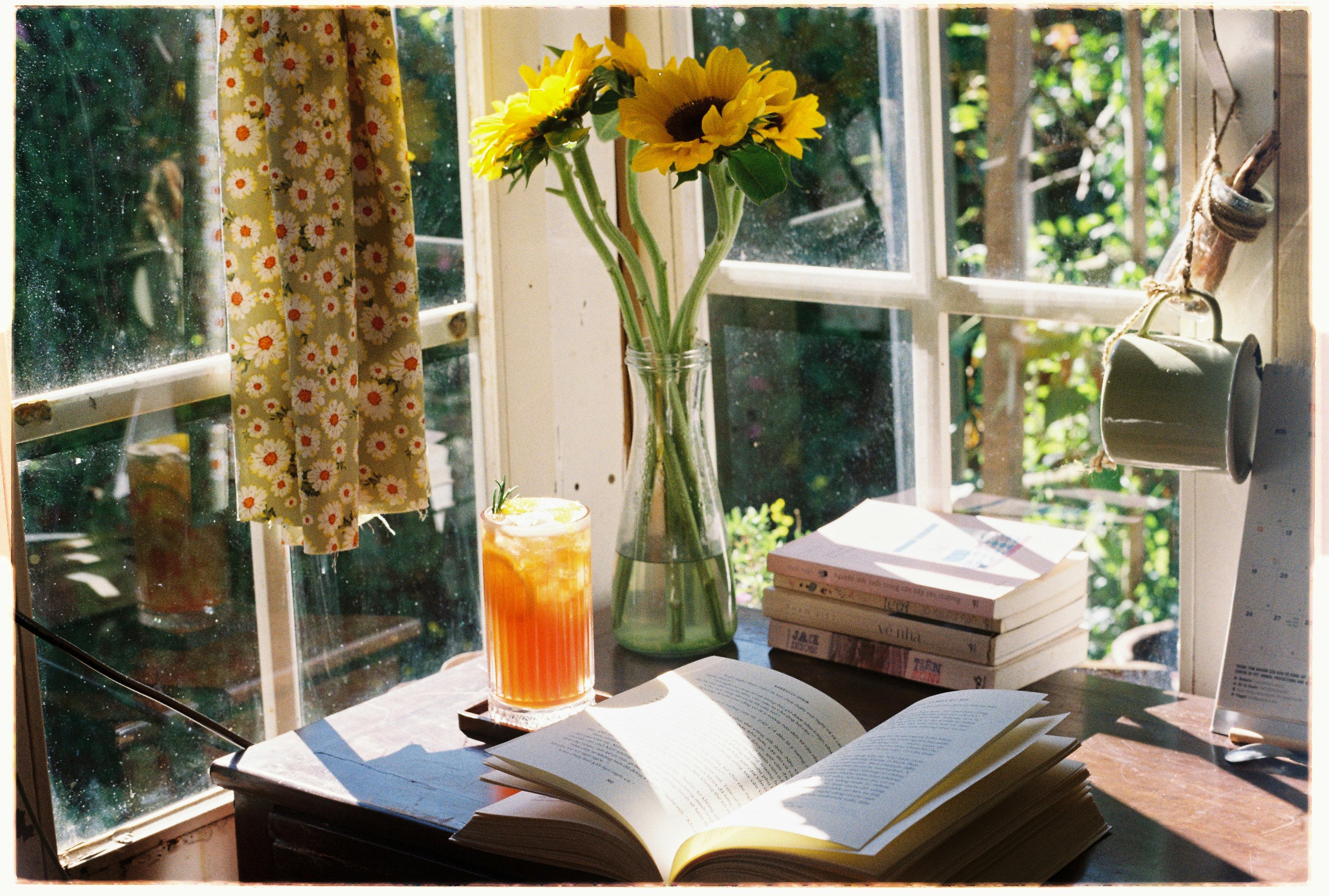 Desk next to a corner of windows, with open book, a cup of iced sweet tea, and a vase of sunflowers