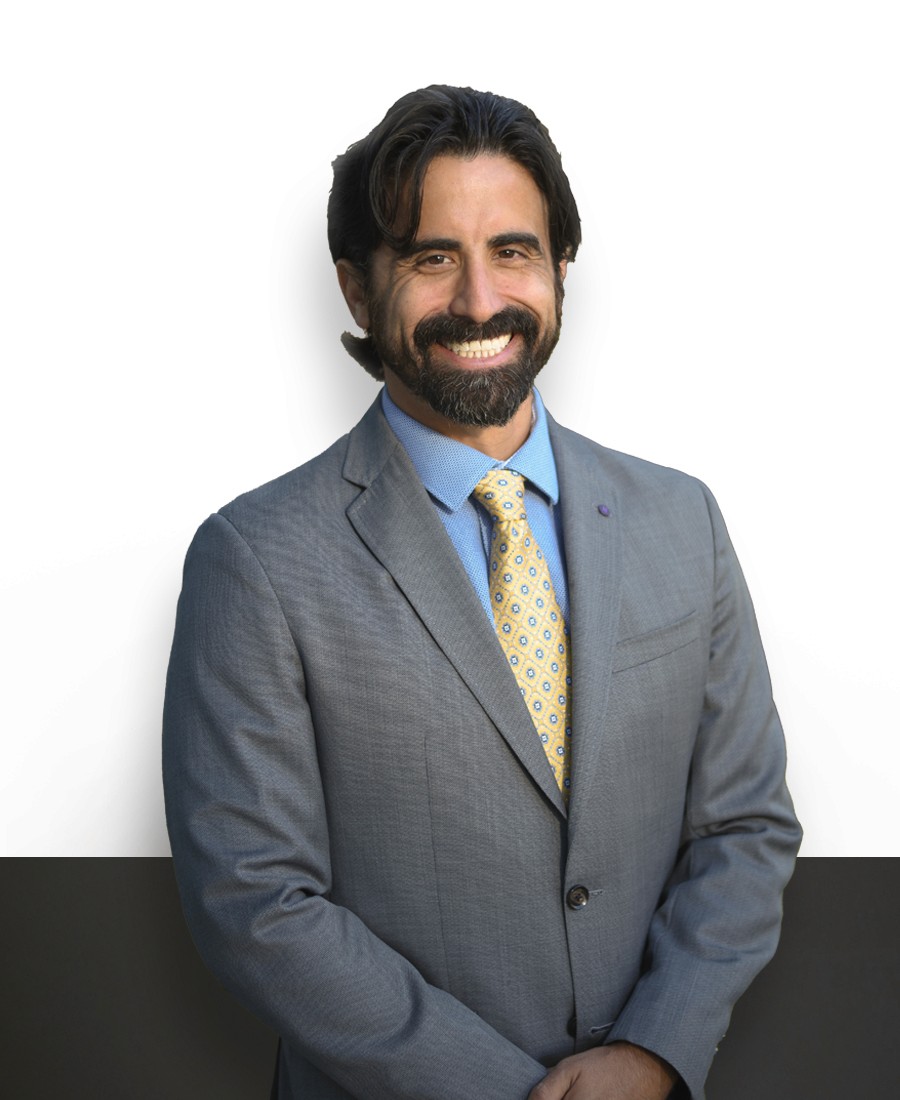 corporate headshot, male with light brown skin and dark styled hair  with a dark beard and mustache, wearing a light grey suit with blue button up shirt and yellow patterned tie, smiling with his hands clasped over each other in a relaxed pose. 