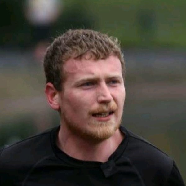 Head-and-shoulders photograph of a person wearing a black top outdoors. The image is tightly cropped on the face and upper torso, with a blurred background and natural lighting.