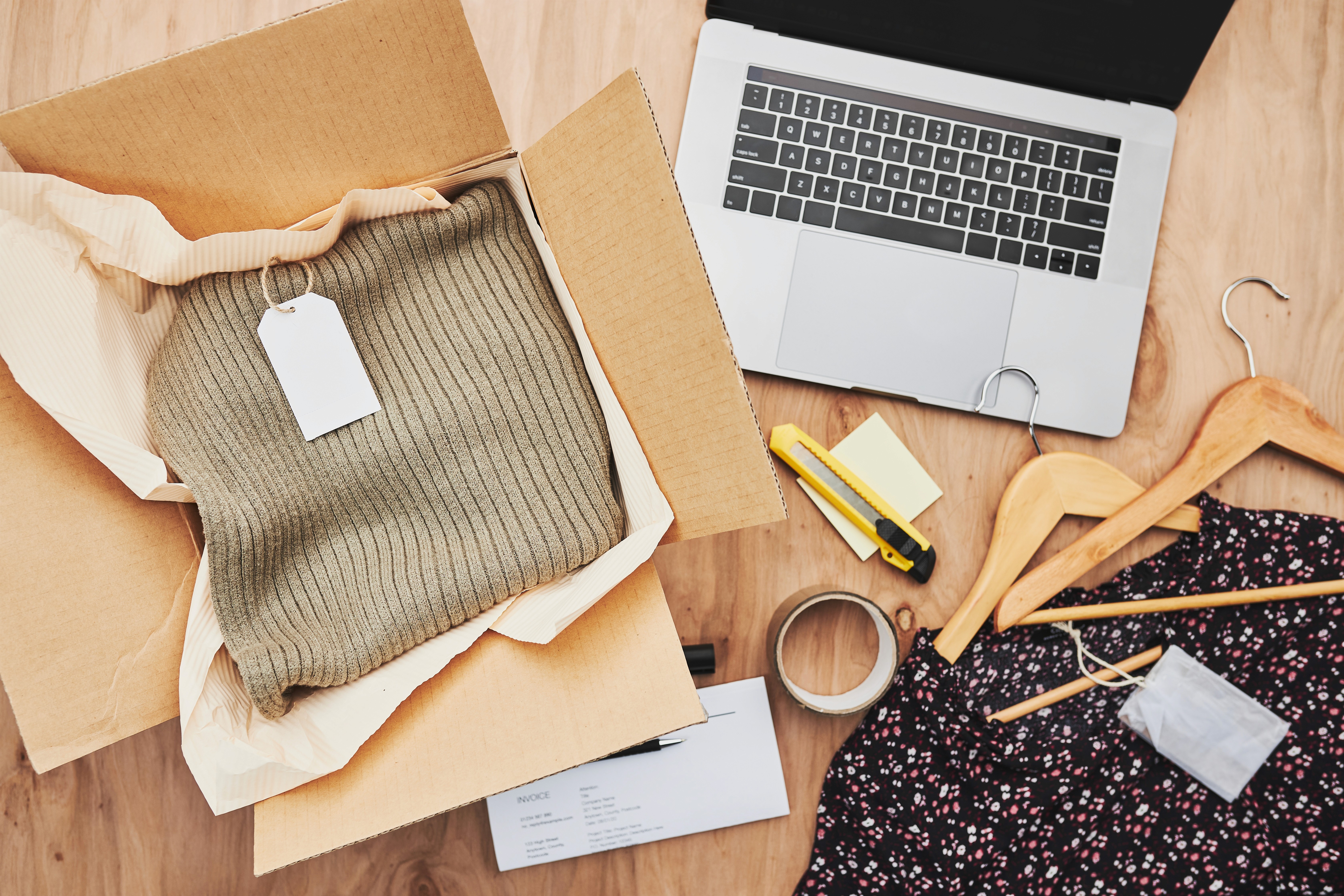 Flat lay of online clothing order being packed with cardboard box, sweater, laptop, and hangers