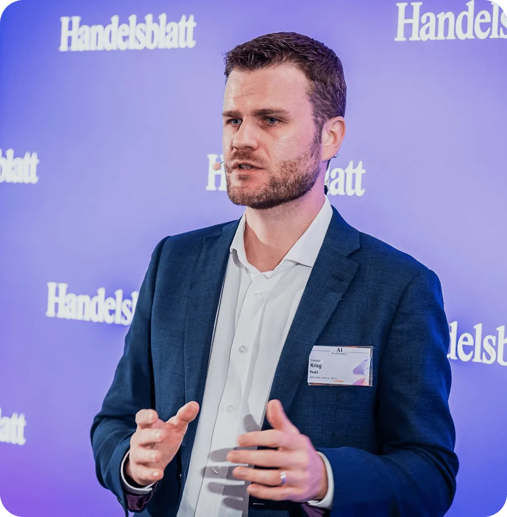 A man in a blazer speaks at an event, with a purple backdrop featuring promotional text.