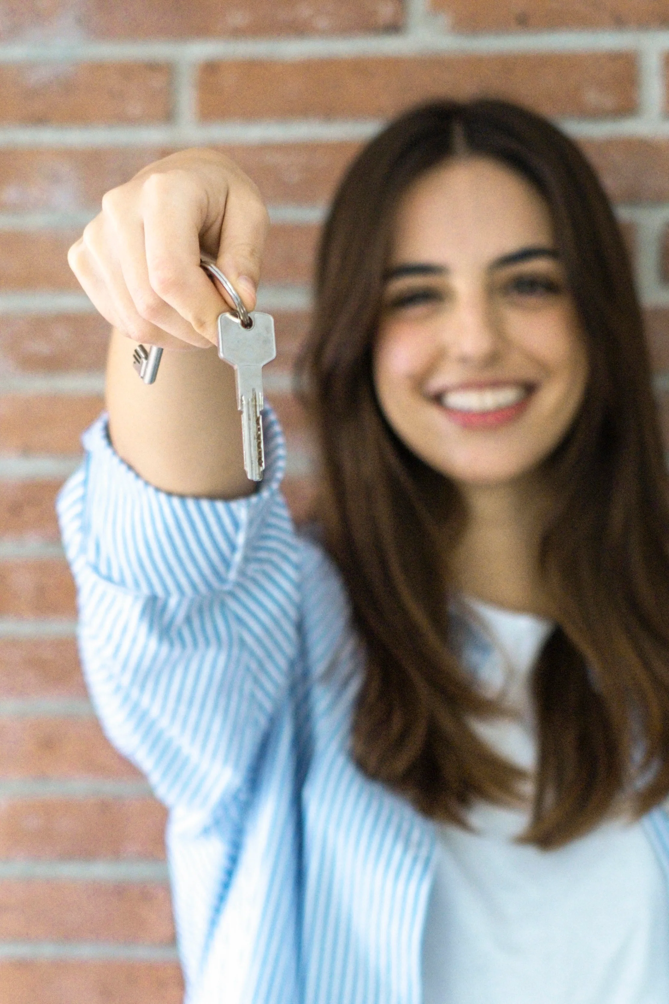 A smiling woman in a blue striped shirt holds up a house key in front of a brick wall background.