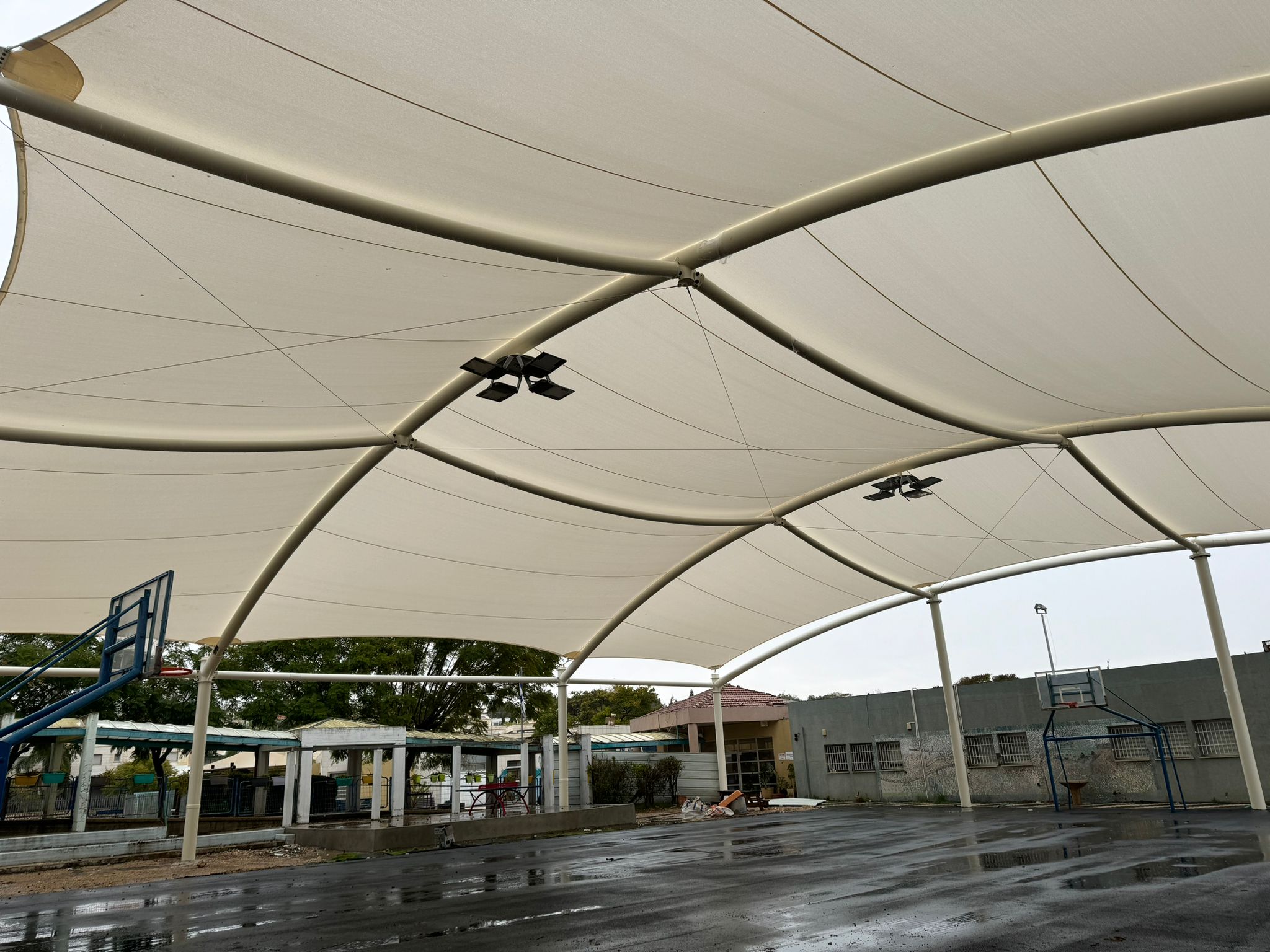 Canopy with curved metal beams and beige fabric roof over a wet, empty sports court with basketball hoop and buildings in the background.