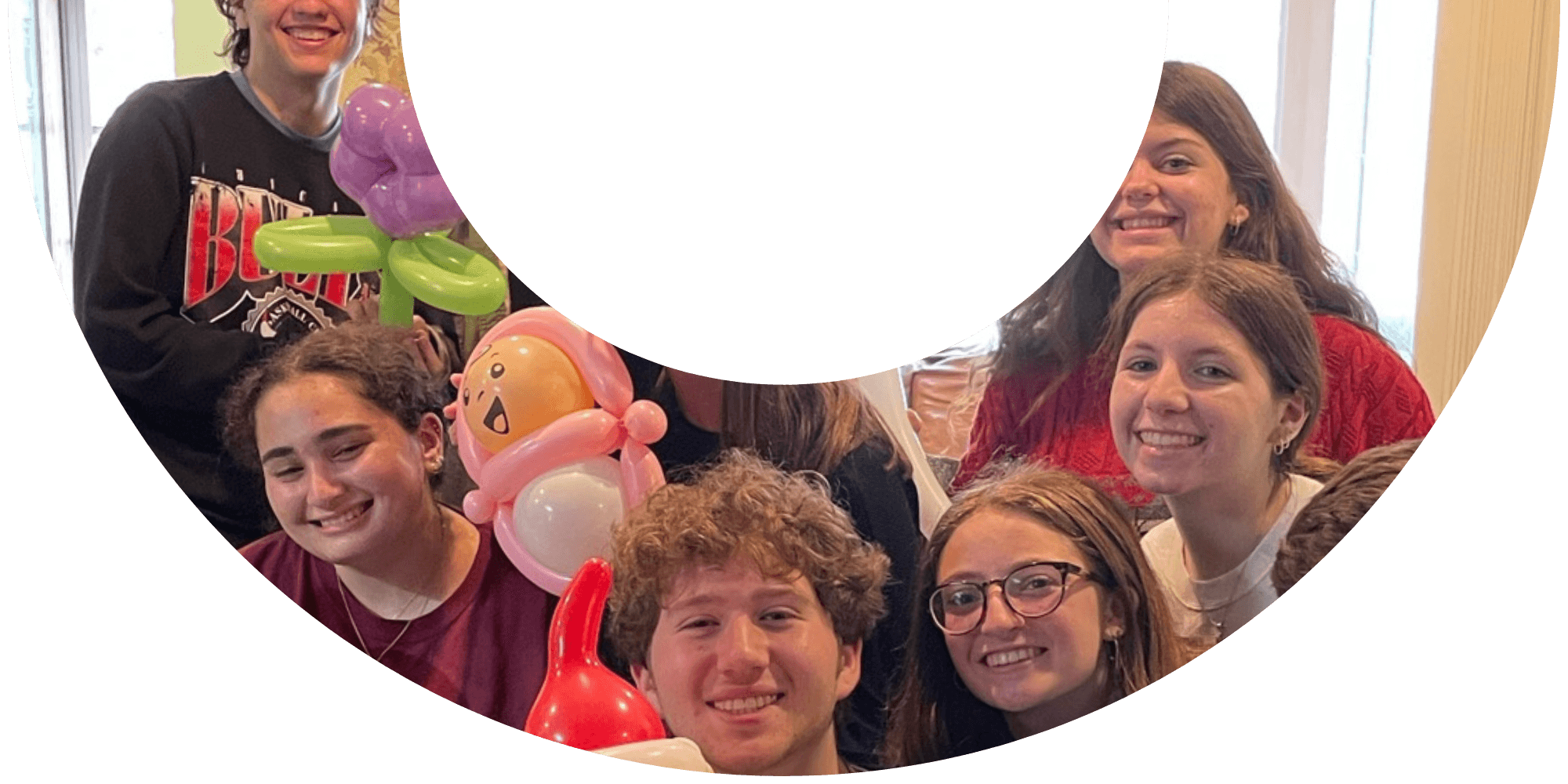 Group of smiling young adults holding balloon art in a brightly lit indoor setting, with a partially cropped smile overlay ontop