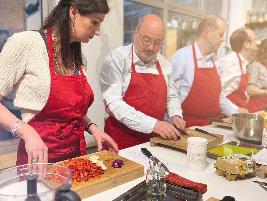 Deux personnes coupant des légumes dans une cuisine lors d’un atelier culinaire, avec d'autres participants en arrière-plan.