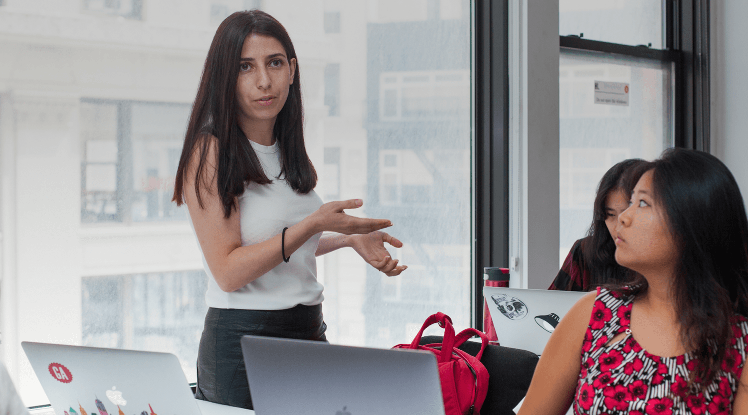 A smiling man with curly hair is sitting at a desk, looking at the camera, with a laptop and a lamp in the background.