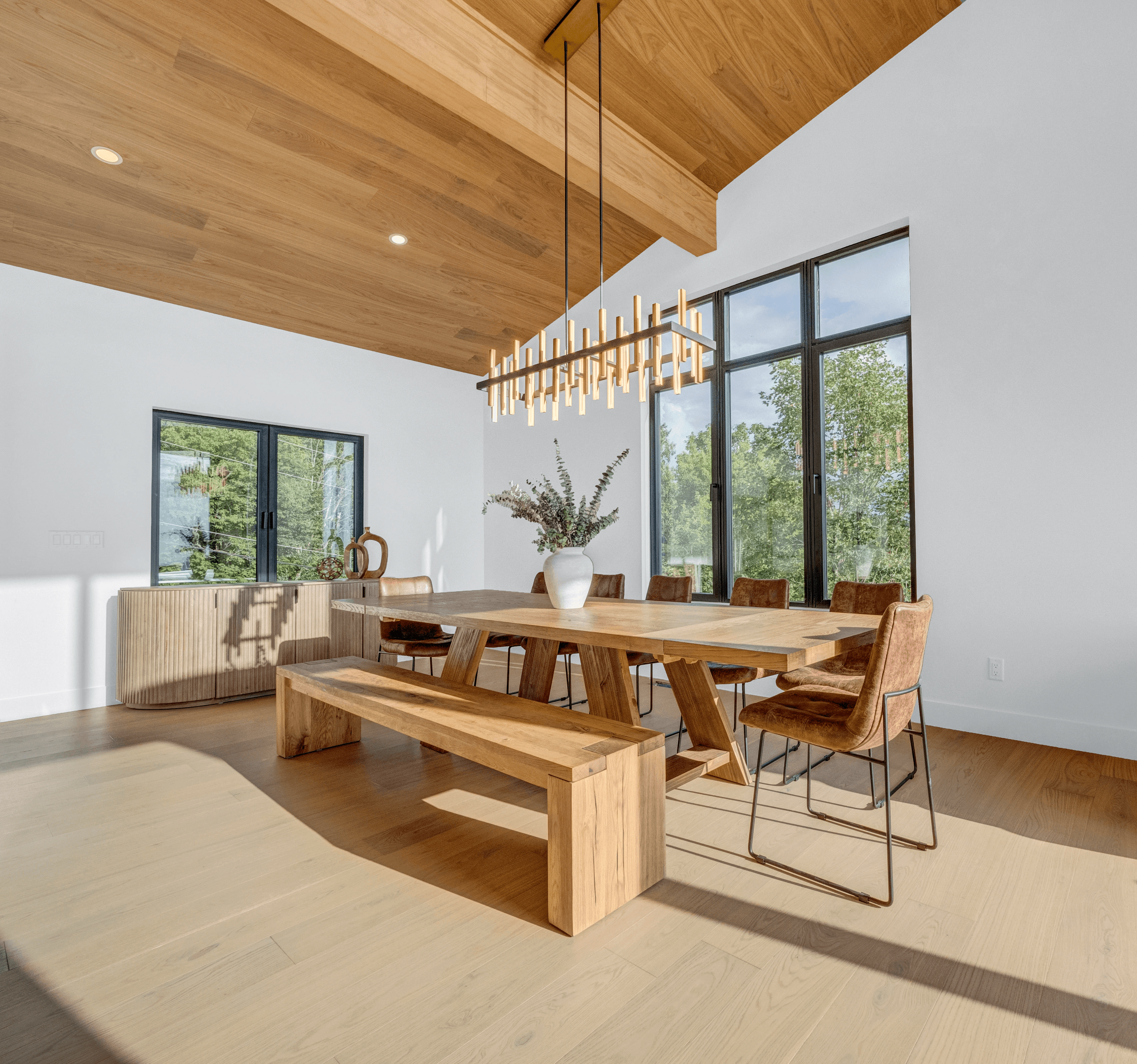 A long natural wood dining table with a modern linear chandelier and leather chairs set against a bright white wall