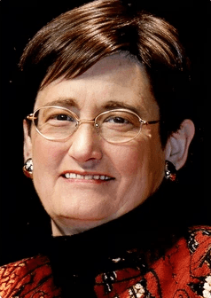 Smiling female therapist with short gray hair, wearing a brown top and a purple shawl, surrounded by a cozy background.