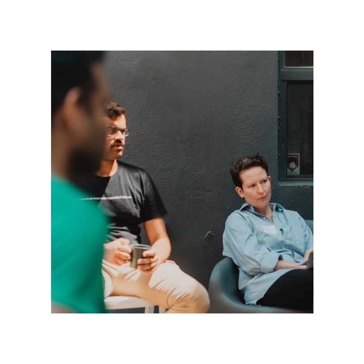 Three people are sitting against a gray wall, engaged in a conversation. One in a black shirt holds a cup, while another in a light blue shirt listens attentively. The atmosphere is casual and focused.